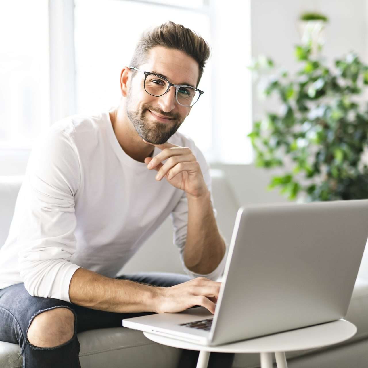 Young attractive guy is browsing at his laptop, sitting at home on the cozy beige sofa at home, wearing casual outfit